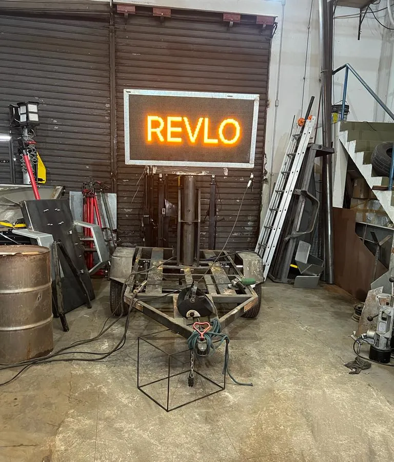 Painel de mensagem variável aceso com a palavra REVLO em LED âmbar, sobre chassi de reboque ainda sem pintura, na oficina em 2023